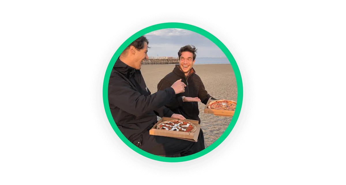 A circular image showing a photo of two people at the beach, holding a pizza box in their hands and looking at each other and smiling